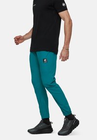 Teal softshell broek met een elastische tailleband, taps toelopende pijpen en een zwart logo-opdruk op de bovenbeen. G gedragen met een zwart T-shirt met korte mouwen en zwarte schoenen.
