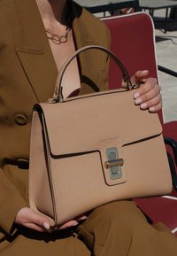 Tan leren handtas met een gestructureerde vorm, minimalistisch ontwerp, contrasterende zwarte randen, gouden hardware en een klep-sluiting met een groene accent.