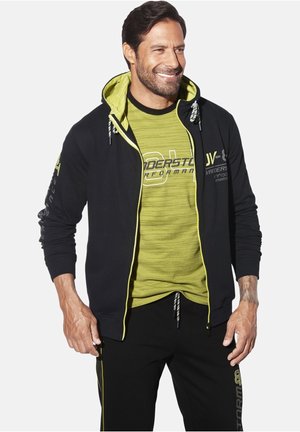 Black zip-up hoodie with a bright green interior and drawstrings, layered over a green striped t-shirt. Black pants with yellow accents.