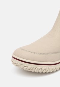 Punta di stivale in pelle beige con cuciture visibili, suola in gomma crema, pattern di battistrada profondo e una sottile striscia rosso scuro sopra la suola.
