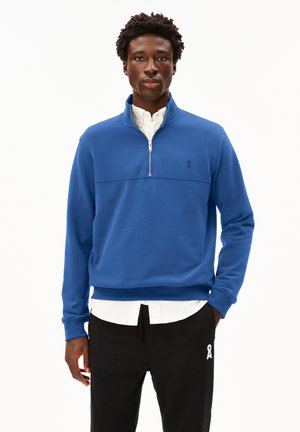Sweatshirt bleu à fermeture quart-zip en tissu doux, avec un ourlet et des poignets côtelés, affichant un logo discret sur la poitrine. Porté sur une chemise blanche.