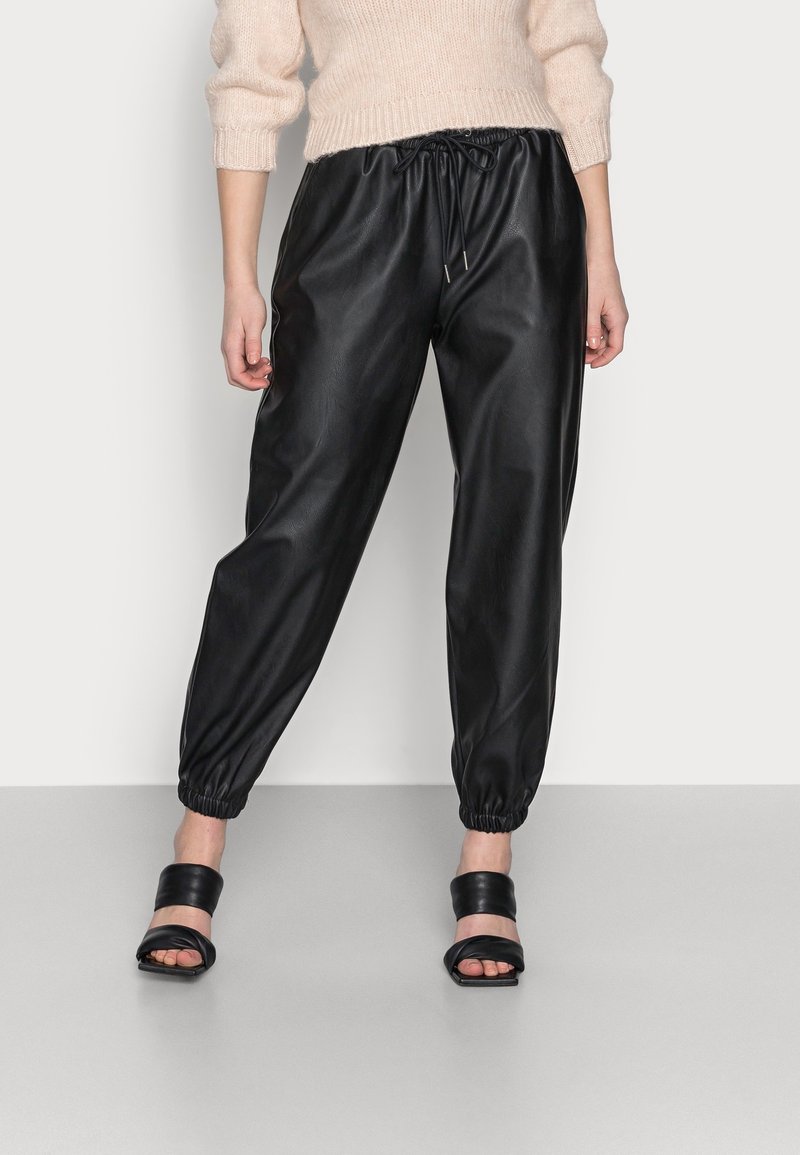 Pantalon jogger en similicuir noir avec taille et chevilles élastiques. Texture lisse, coupe fuselée. Porté avec des sandales slides noires.