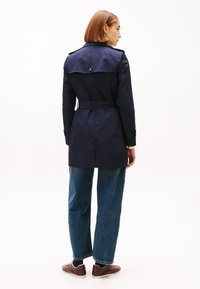 Kvinne iført en marineblå trenchcoat med belte, blå jeans og burgunderrøde joggesko, stående med ryggen mot, foran en hvit bakgrunn.