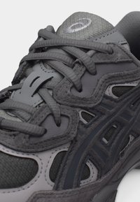 Gros plan sur une chaussure de sport gris foncé montrant un tissu en mesh, des empiècements en daim et des lacets croisés sur la languette avec un logo cousu.