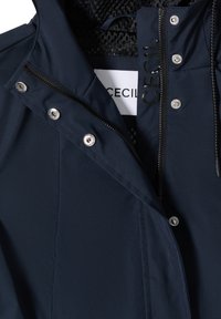 Veste bleu marine avec fermeture éclair et boutons-pression, partiellement ouverte pour révéler une doublure en filet noir et une étiquette blanche portant le texte "CECIL" à l'intérieur.
