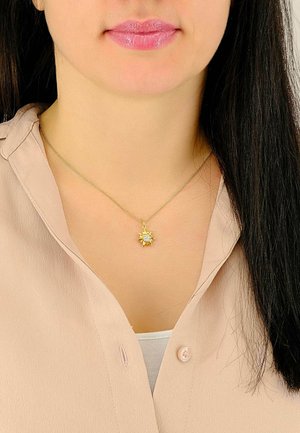 Collier en chaîne dorée avec un pendentif en forme de soleil et une pierre blanche centrale, porté sur un blouse beige clair.