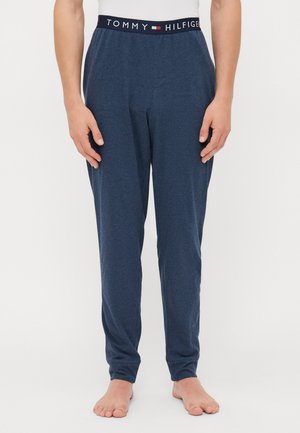 Mann steht barfuß, trägt marineblaue Tommy Hilfiger Loungehosen mit Logo-Bund und ein weißes Oberteil vor einem weißen Hintergrund.