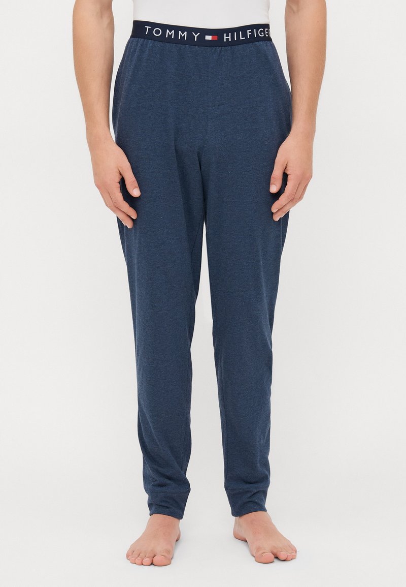 Man die blote voeten heeft en navy blauwe loungebroeken van Tommy Hilfiger draagt met een logo tailleband en een witte top, tegen een witte achtergrond.