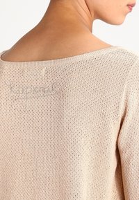 Ljusbeige stickad tröja med ett löst, brett halsringning och ett texturerat mönster, med subtil märkning sydd nära axeln.