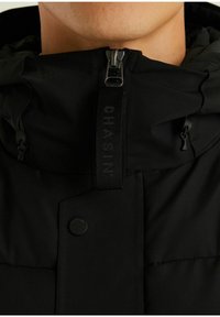 Primer plano de una persona que lleva una chaqueta negra con capucha, cerrada hasta el cuello con una lengüeta de cremallera de marca y un botón a presión visibles.