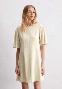 Kurzärmliges, knielanges Kleid aus weichem Creme-Stoff. Hat einen rundem Ausschnitt, eine gerade Silhouette und subtile vertikale Nähte.