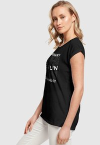 Sort t-shirt med rund halsudskæring og opsmøgte ærmer, der har et hvidt grafisk design med teksten "Berlin" og "Tyskland."