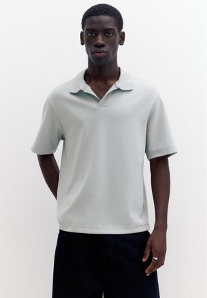 Junger Mann mit dunkler Haut, der ein hellgraues Polohemd mit kurzen Ärmeln und dunkle Hosen trägt und vor einem schlichten weißen Hintergrund steht.