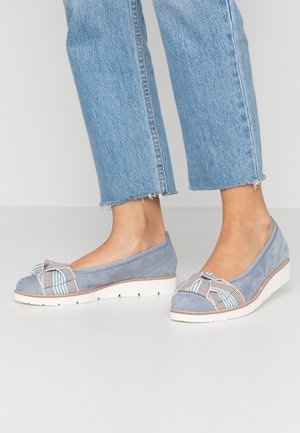Kvinde iført lyseblå ruskind slip-on sko med hvide såler og stribede stofsløjfer, kombineret med blå jeans med frynset kant.
