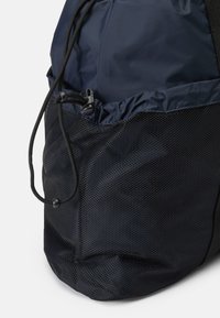 Sötétkék hátizsák húzózsinórral, hálós elülső zsebbel és sima nylon textúrával, a funkcionalitásra és a tartósságra tervezve.