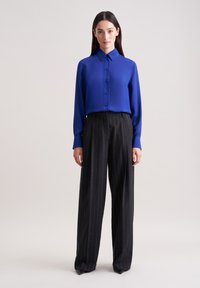 Blaue Bluse mit Kragen und Frontknöpfen, kombiniert mit schwarzen, gestreiften, weit geschnittenen Hosen. Glatte Stoffqualität und strukturierte Passform.