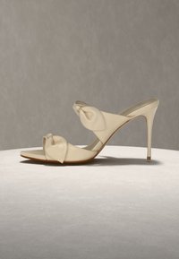 Μπεζ mule με ψηλό τακούνι, δύο λουριά με κόμπους, μυτερό τακούνι στιλέτο και ανοιχτό δάχτυλο πάνω σε ανοιχτή επιφάνεια και γκρι φόντο.
