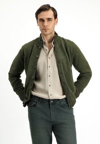 Green knit cardigan with a ribbed texture, button closure, and high collar, worn over a light beige button-up shirt and dark jeans.