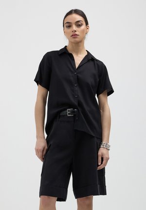 Donna che indossa una camicia nera a maniche corte con bottoni, pantaloni corti neri con cintura e tasche, e un bracciale d'argento spesso, in piedi contro uno sfondo neutro.