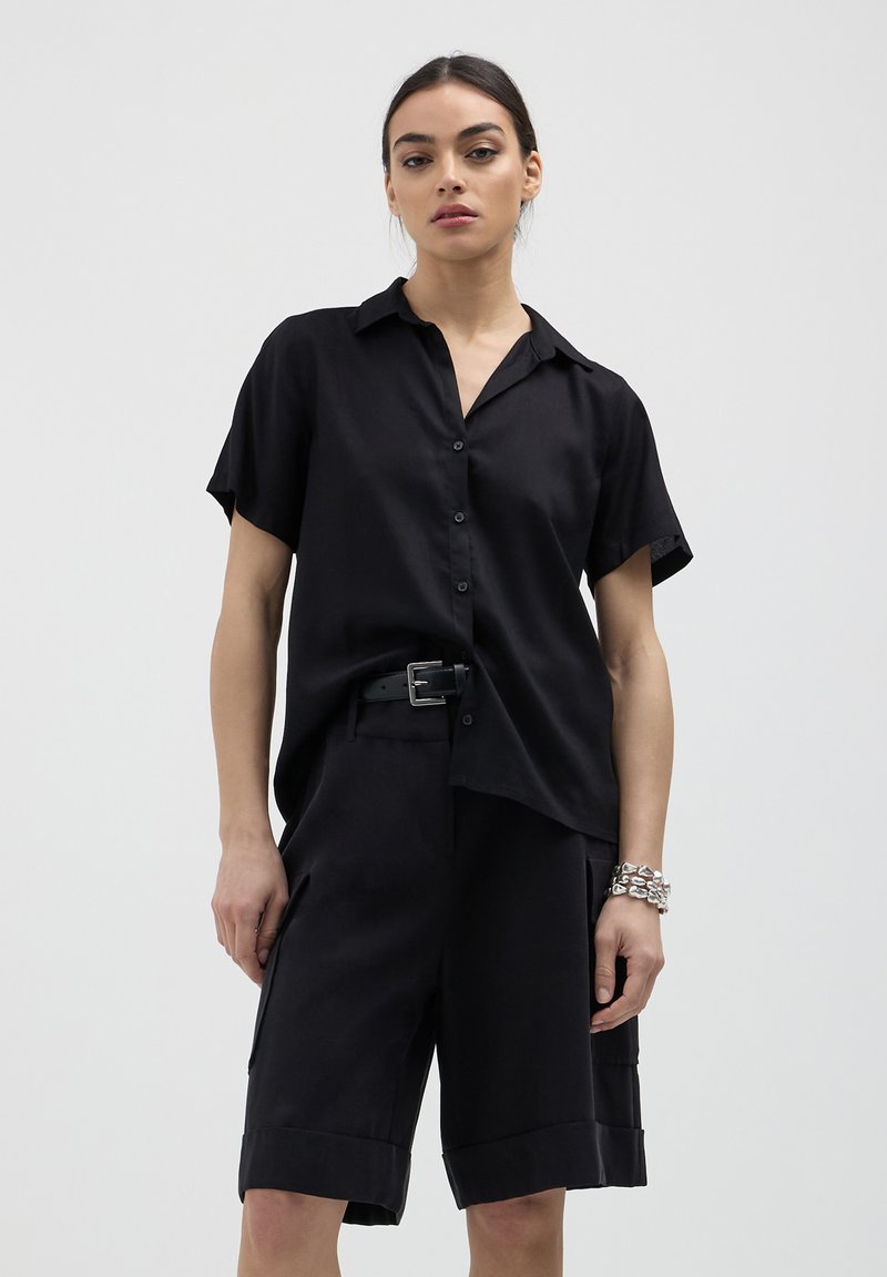 Femme portant une chemise à manches courtes noire à boutons, un short noir ceinturé avec poches et un bracelet en argent massif, se tenant contre un fond uni.