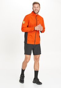 Orange letvægtsjakke med sorte sidelommer, lynlås fortil og en lille gul lomme detalje. Matchet med sorte shorts og atletiske sko.
