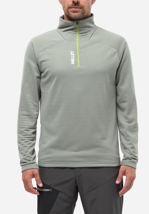 Homme portant un pull quart-de-zip côtelé gris avec une fermeture éclair jaune et un pantalon gris foncé avec une poche zippée, debout face à la caméra.