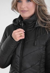 Schwarze isolierte Jacke mit durchgehendem Reißverschluss und verstellbarer Kapuze. Verfügt über ein gestepptes Design und eine weiche, glänzende Textur. Hand greift nach dem Reißverschluss.