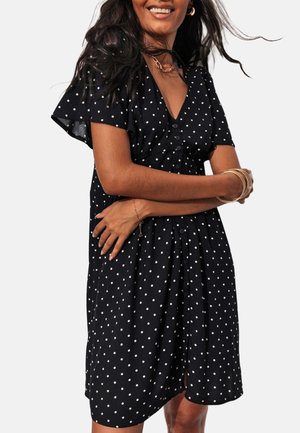 Femme souriante portant une robe noire à pois blancs, manches courtes et boutons sur le devant, bras croisés sur la taille.