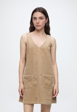 Vrouw met lang bruin haar die een mouwloze beige corduroy jurk met zakken aan de voorkant draagt, staand tegen een effen lichte achtergrond.