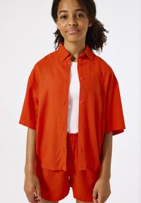 Camisa corta de lino roja con mangas cortas y botones en la parte delantera, llevada sobre una camiseta blanca de canalé, combinada con pantalones cortos rojos a juego.