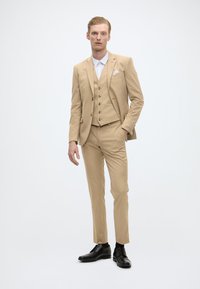 Costum bej format trei piese, cu sacou ajustat, vestă și pantaloni personalizați. Cămașă albă cu guler. Pantofi eleganți negri. Textură netedă.