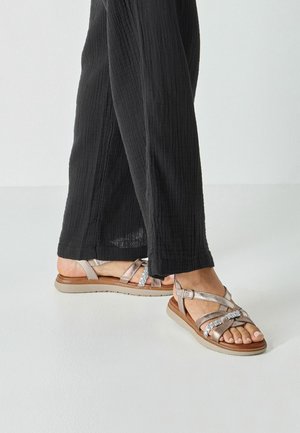 Sandały na paski w kolorze srebrnym metalicznym i beżowym, z krzyżującymi się paskami, regulowaną klamrą oraz jasnobrązową gumową podeszwą.