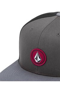 Grå tygmössa med svart topp, med en cirkulär vinröd lapp med en vit logotyp i mitten. Skärmen är platt med texturerad sömnad.