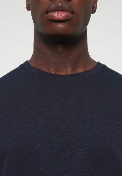 Close-up of a person’s lower face, neck, and upper chest wearing a dark blue crewneck shirt against a plain background.
