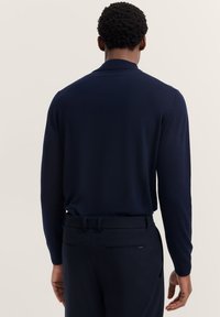 Marinefarbener langärmeliger Pullover mit flachem Kragen, schmaler Passform, glatter Textur und gerippten Bündchen, kombiniert mit dunklen, maßgeschneiderten Hosen.