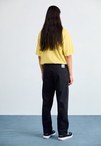 Gul bomullströja med korta ärmar och avslappnad passform, matchad med svarta byxor och svarta sneakers, sedd bakifrån.