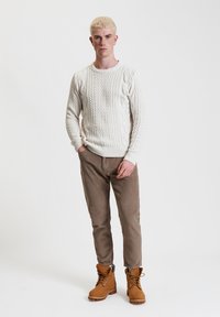 Maglione crema lavorato a maglia con motivo a treccia, abbinato a pantaloni in corduroy marroni e robuste scarpe stringate color nocciola, in piedi su uno sfondo bianco.