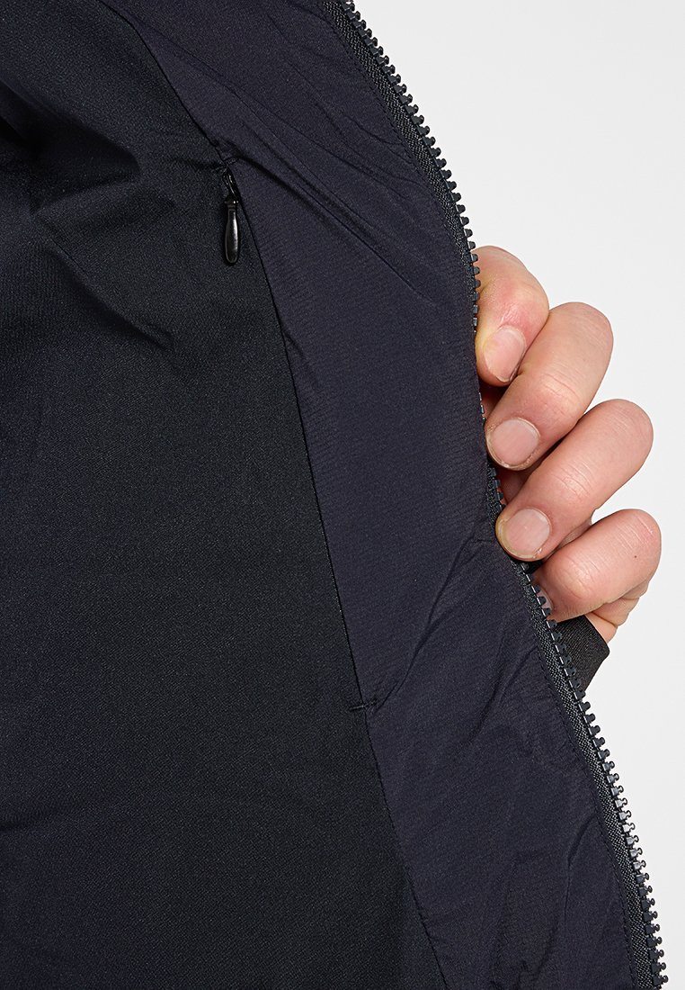 Intérieur d'une veste zippée noire présentant un tissu lisse et léger au texture plate, montrant une main tenant le bord près d'une poche latérale.