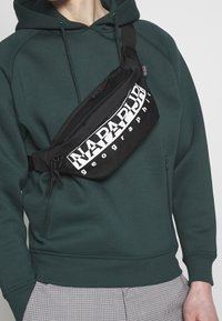 Černá nylonová ledvinka s výrazným bílým logem "NAPAPIR" a zipovým uzávěrem, nošena přes tělo na zelené mikině.