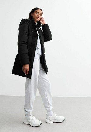 Zwarte pufferjas met capuchon, rits aan de voorkant en ritszakken, gedragen over een lichtgrijze sweatshirt en sweatpants, gecombineerd met witte sneakers.