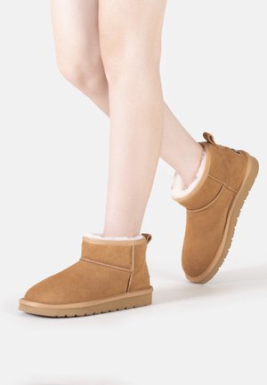 Tan suede ankle boots with a soft white shearling lining and rubber sole. The design features minimal stitching and a pull tab at the back.