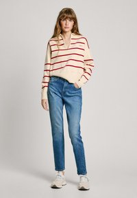 Maglione a righe crema con linee rosse, collo a V e tasca, abbinato a jeans azzurri chiari e sneakers beige, che mostra un look casual.