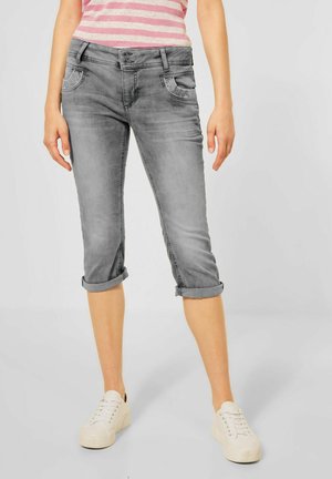 Femme portant un jean gris retroussé et court, des baskets blanches, et une chemise rayée rose et blanche, debout devant un fond uni.