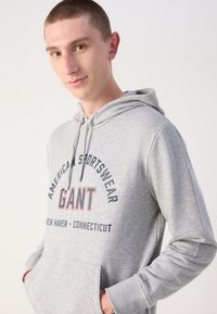 Sudadera con capucha gris hecha de una mezcla de algodón, con un bolsillo frontal y texto impreso, que presenta cordones blancos y puños acanalados.
