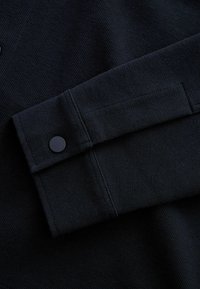Gros plan d’un poignet de manche en tissu noir avec un bouton-pression noir et des détails de couture visibles.