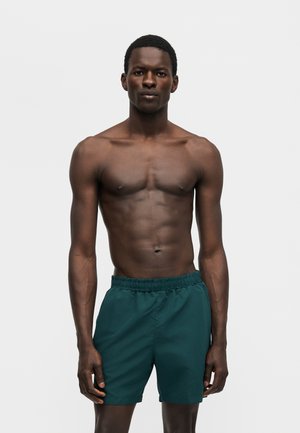 Γυμνασμένος άνδρας με σκουρόχρωμο δέρμα, φορώντας σκούρα πράσινα μαγιό, στέκεται μπροστά σε μονόχρωμο ανοιχτό γκρι φόντο.