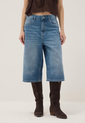 Frau trägt kniehohe blaue Jeansshorts, kombiniert mit dunkelbraunen kniehohen Wildlederstiefeln und einem kurzen dunkelbraunen Oberteil.