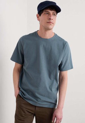 Kurzärmeliges T-Shirt mit Rundhalsausschnitt aus gedämpftem Tencel in Teal. Kombiniert mit braunen Hosen. Das Model trägt eine marineblaue Mütze. Einfaches, lässiges Design.