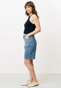 Marineblaue gerippte Tanktop, hoch taillierter blauer Denimrock mit fransigem Saum und hellen Schuhen, vor einem einfarbigen Hintergrund.