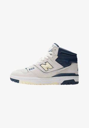 High-top sneaker in off-white suede with navy accents. Features perforated toe box, padded collar, and thick rubber sole with contrasting tread.